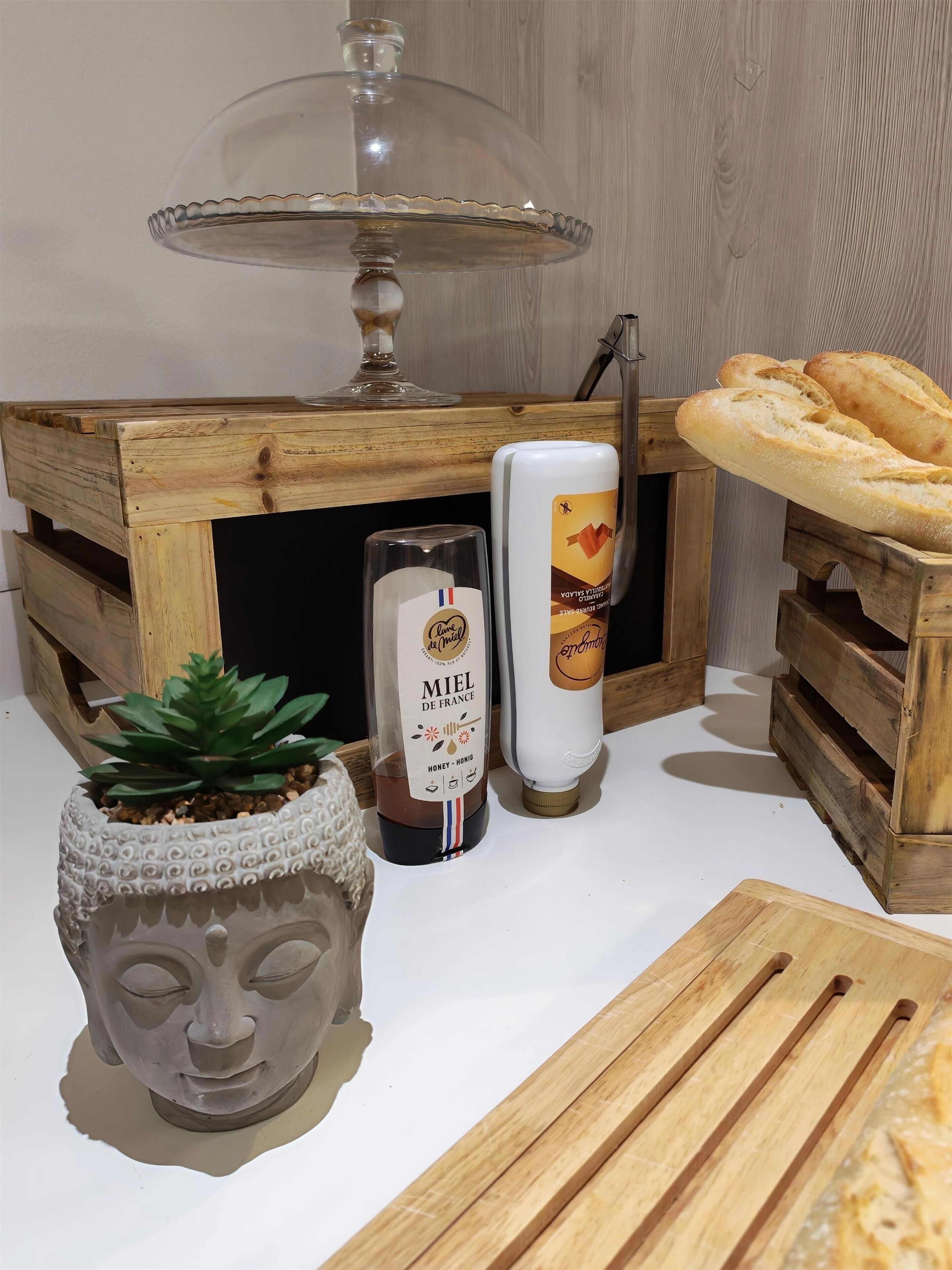 A wooden table with a glass dome lamp, a bottle of Miel de France honey, a bottle of sparkling water, a stone face sculpture, and a wooden cutting board.