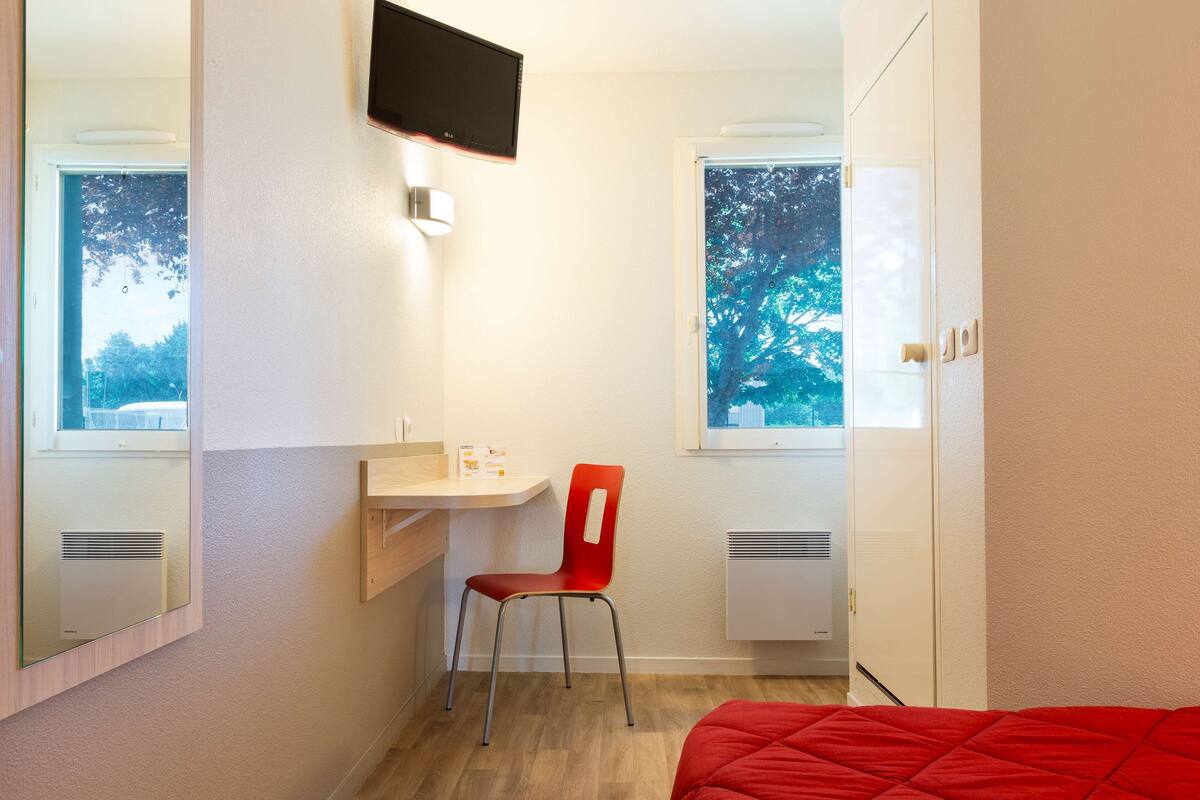 A compact hotel room with a single bed, a desk with a chair, a TV mounted on the wall, and a window with a view of trees.
