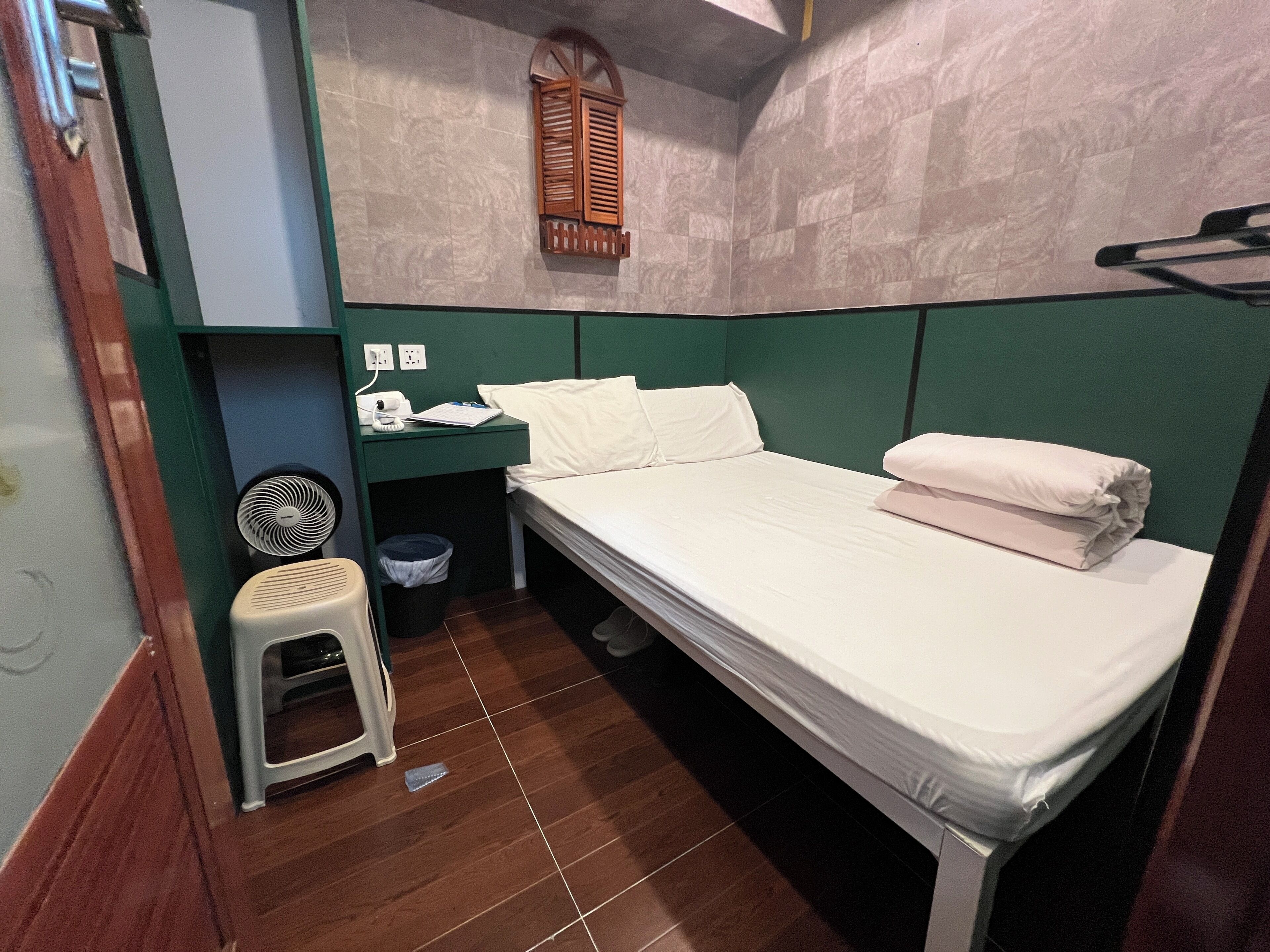 A small, compact room with a single bed, a small stool, a fan, and a window with shutters.