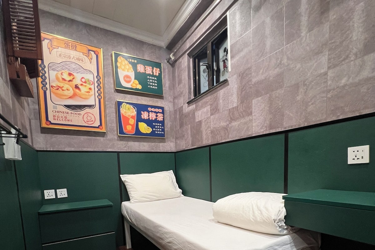 A small, neatly arranged room with a single bed, a green headboard, a small shelf, and a pair of shoes on the floor.