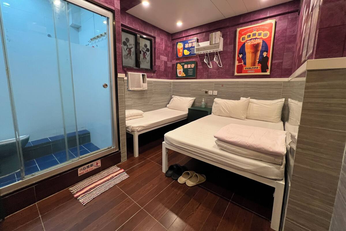A hotel room with two single beds, a glass door, a small air conditioning unit, and a poster on the wall.