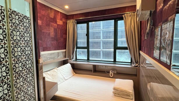 A compact hotel room with a single bed, a small desk, a window with curtains, and a decorative panel on the left. - Venetian Hostel (Kowloon)