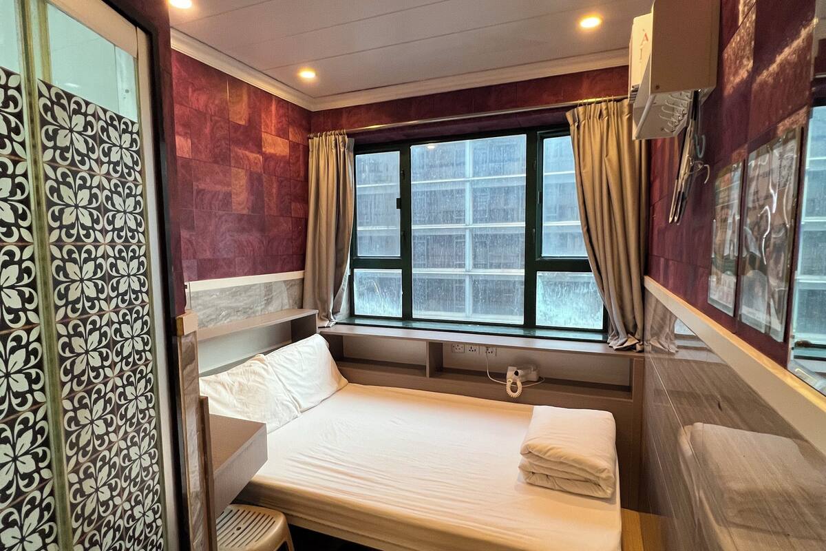 A compact hotel room with a single bed, a small desk, a window with curtains, and a decorative panel on the left.