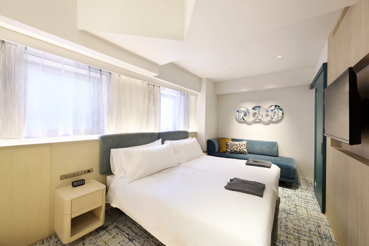 Habitación de hotel con una cama grande, una mesita de noche con un reloj, un sofá, un televisor montado en la pared y una ventana con cortinas traslúcidas.