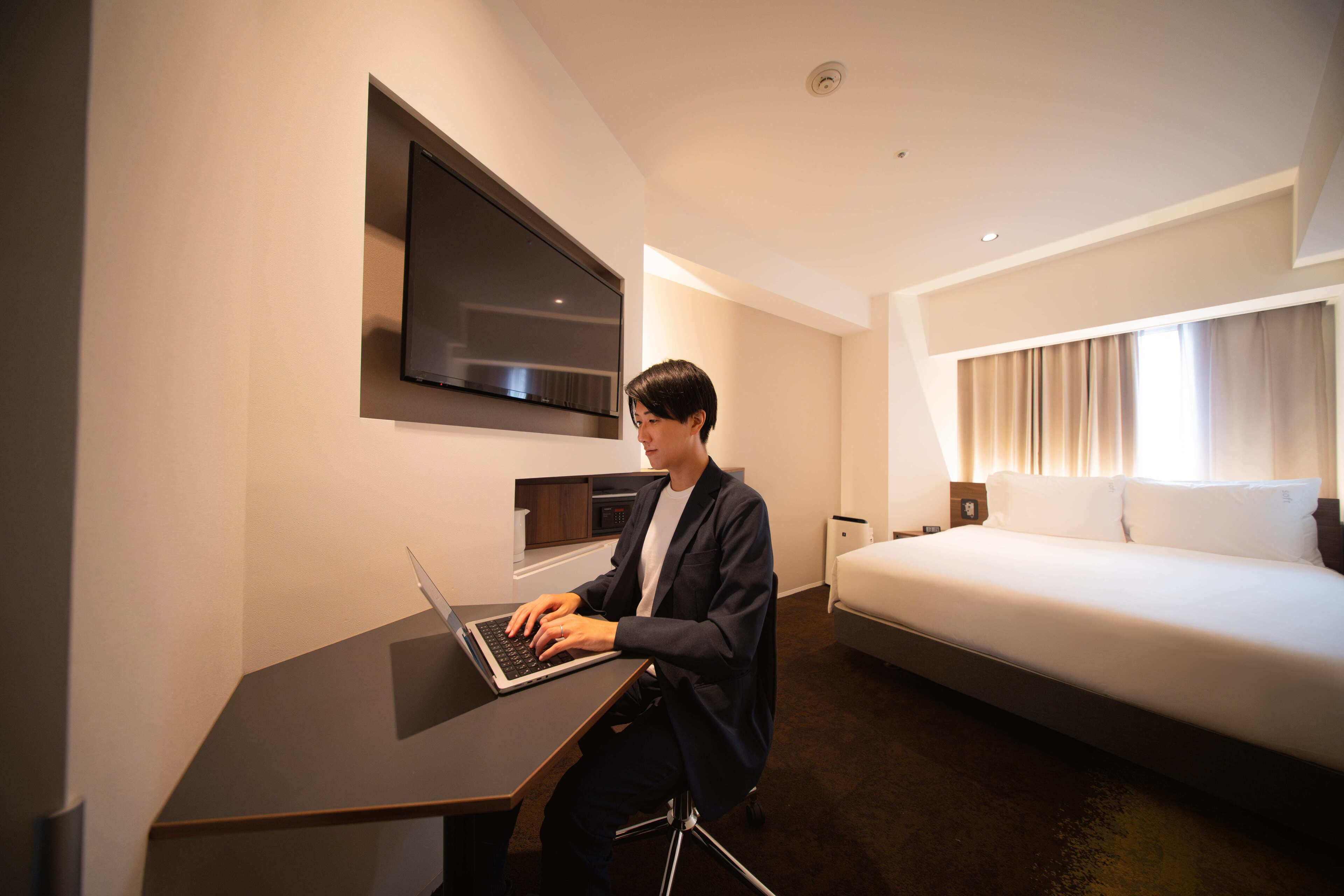 Ein Hotelzimmer mit einem Bett, einem Schreibtisch mit einem Laptop, einem Flachbildfernseher und einem Fenster mit Vorhängen.