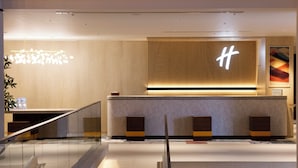 A modern hotel reception area with a wooden reception desk, a large illuminated 'H' sign, and a glass railing.