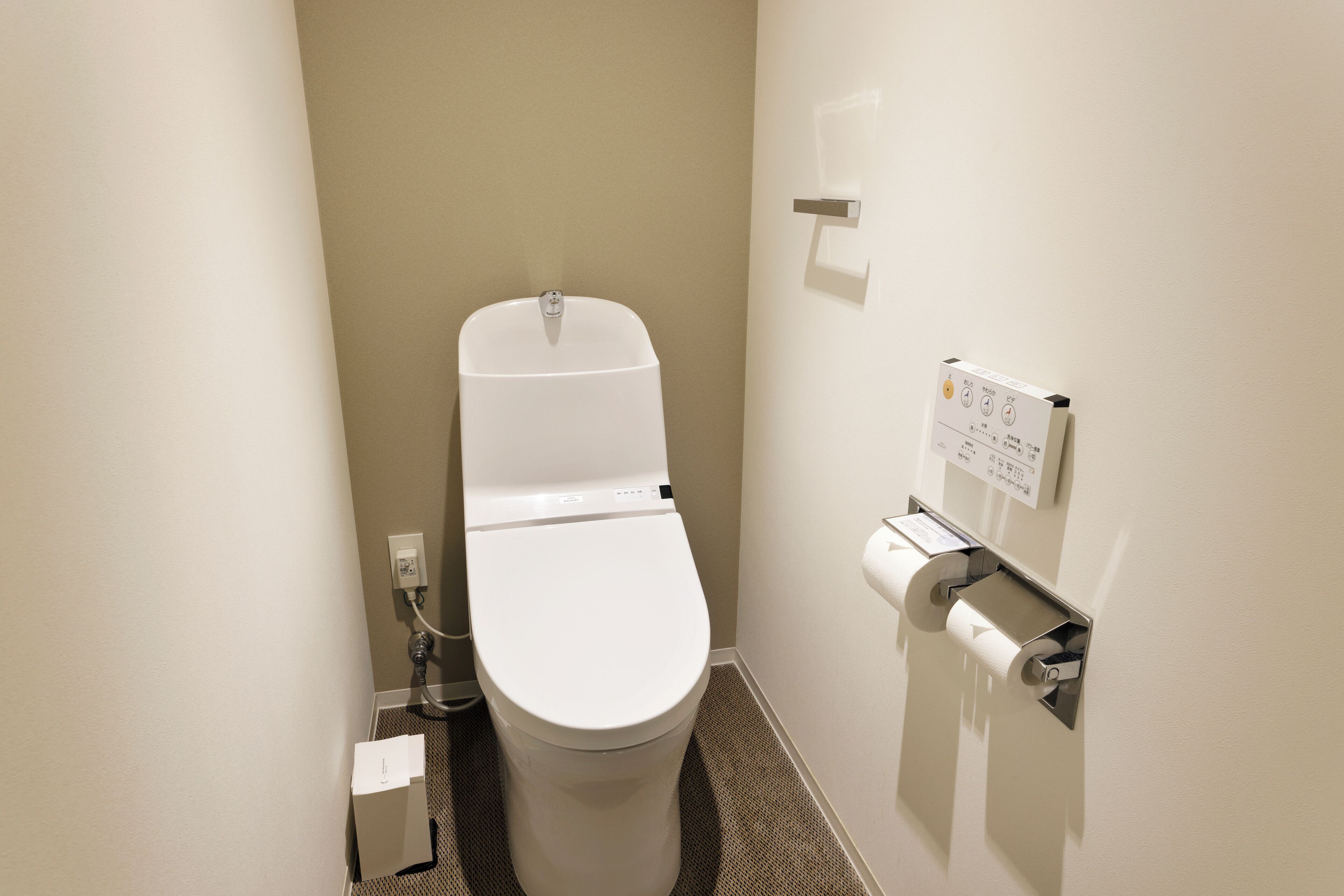 Une petite salle de bain moderne, avec des toilettes blanches, un distributeur d’essuie-tout et un panneau de commande pour la chasse d’eau fixé au mur.