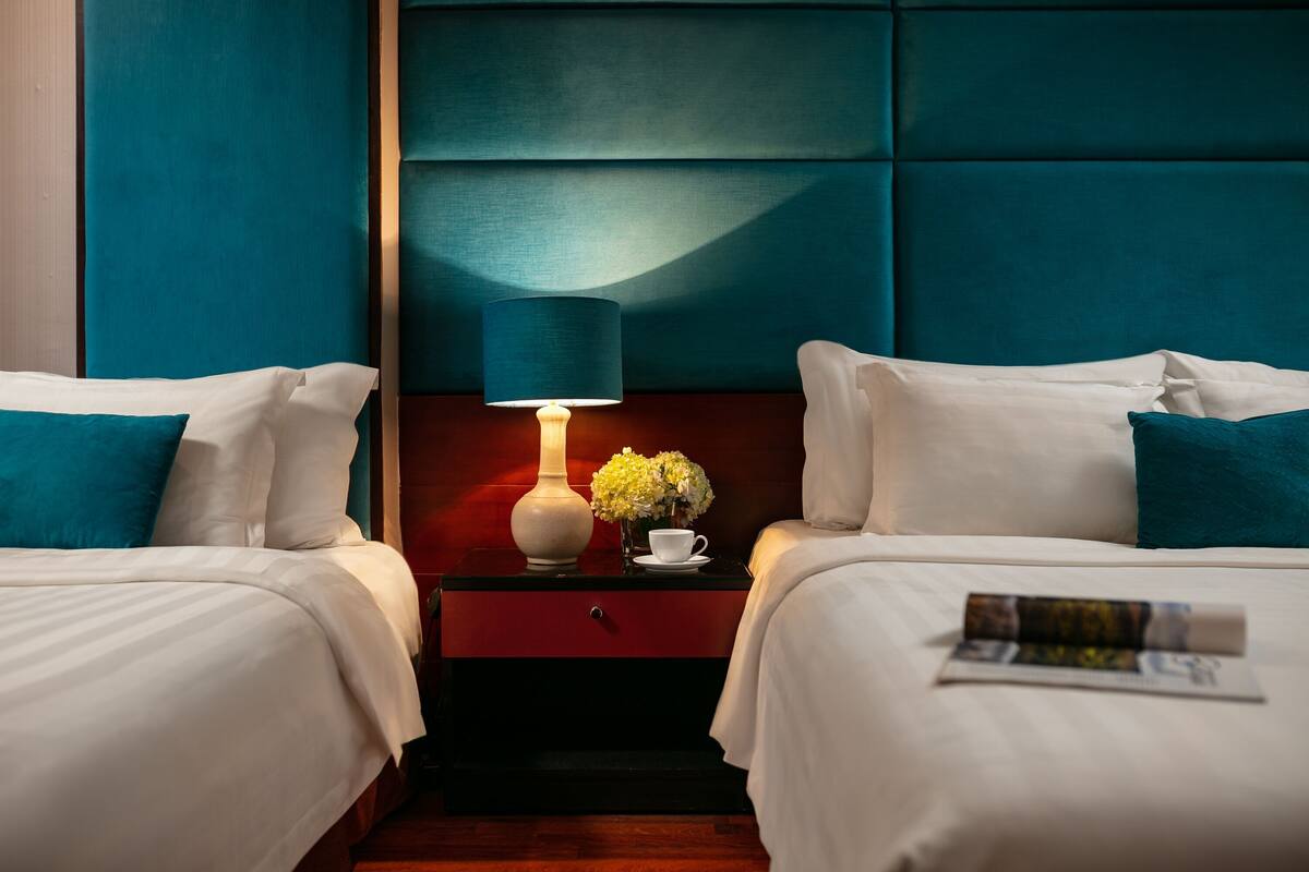 Quarto de hotel com duas camas, uma mesinha de cabeceira com abajur, um vaso com flores e uma revista sobre a cama.