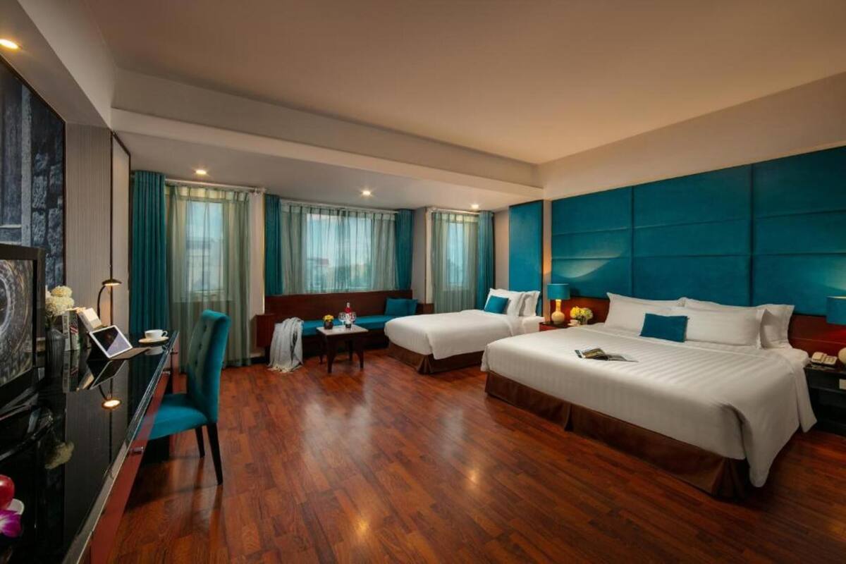 Quarto de hotel com duas camas, uma escrivaninha, uma cadeira, um sofá e uma televisão.