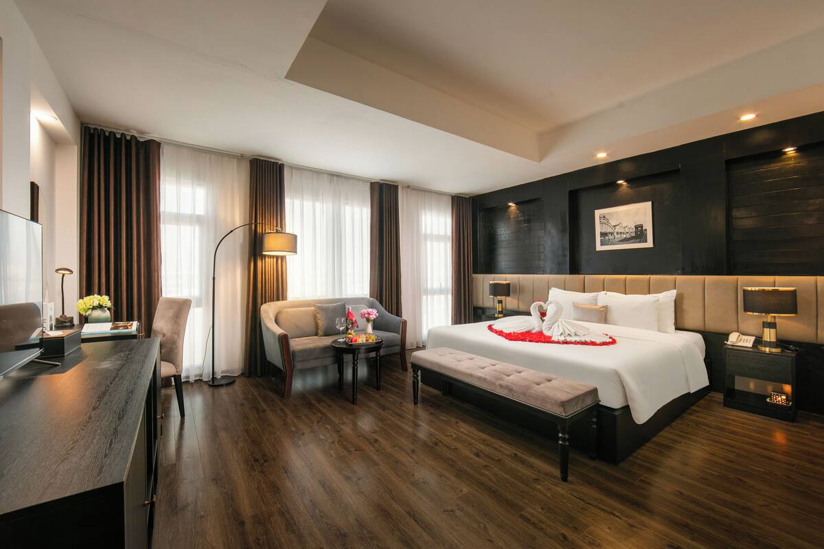 Quarto de hotel moderno com uma cama grande, uma escrivaninha, uma cadeira, uma mesa pequena e uma mesinha de cabeceira com luminária.