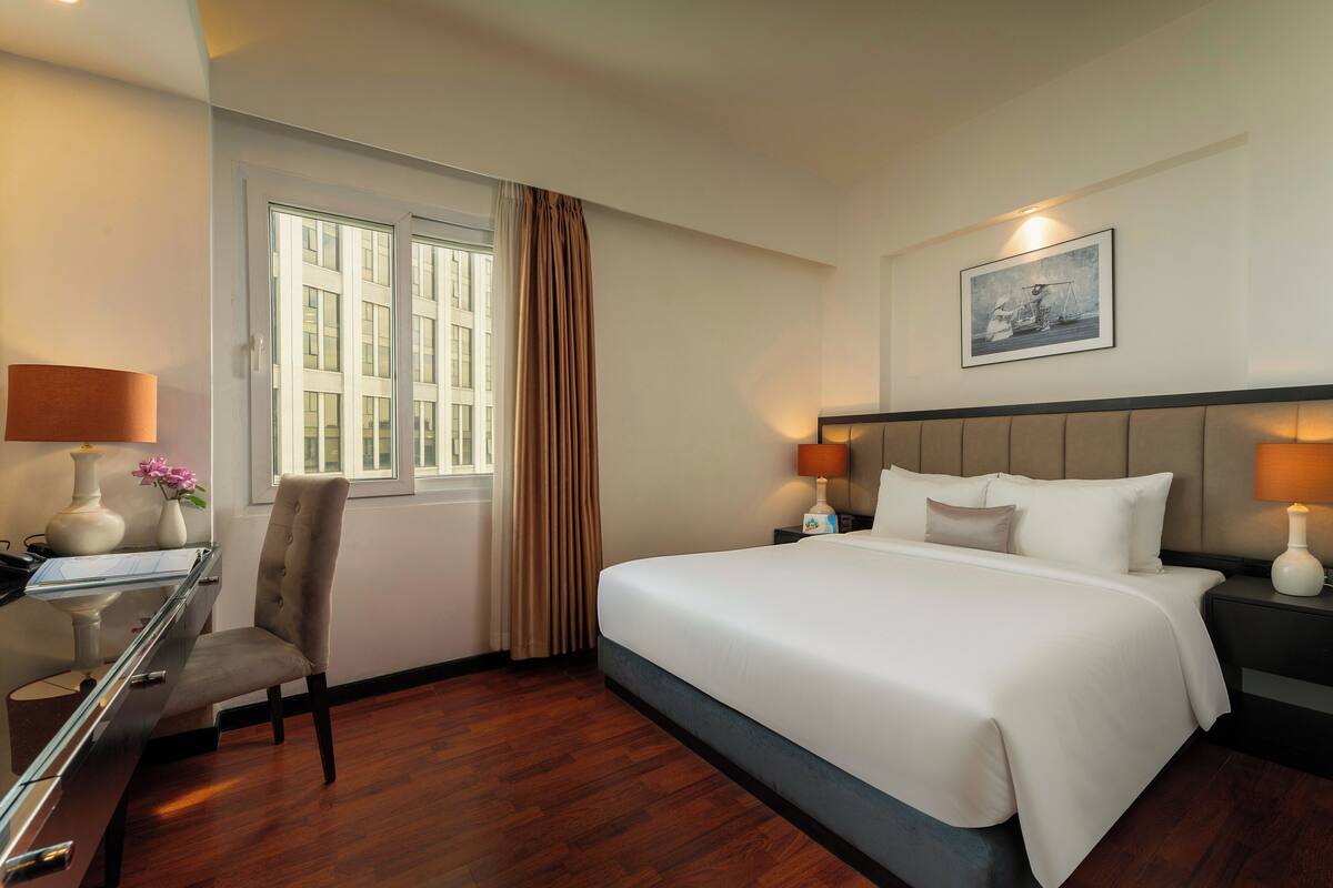 Quarto de hotel com uma cama grande, uma escrivaninha com luminária, uma cadeira e uma janela com cortinas.