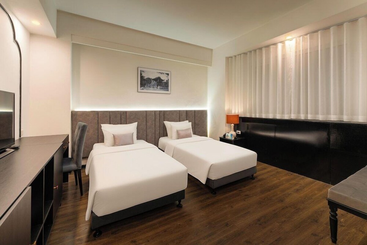 A hotel room with two beds, a desk, a chair, a TV, and a window with blinds.