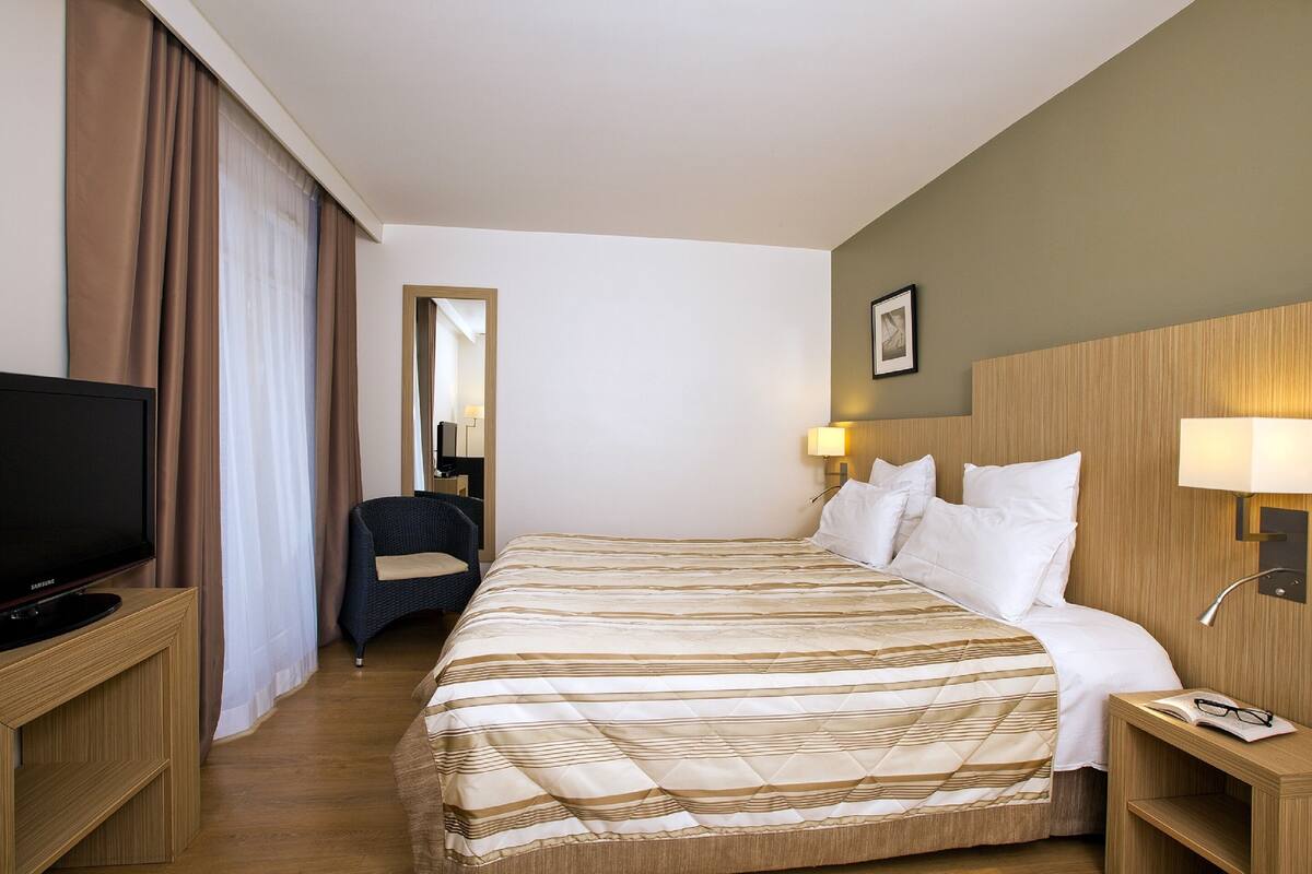 Habitación de hotel con cama, televisión, sillón y escritorio con lámpara.