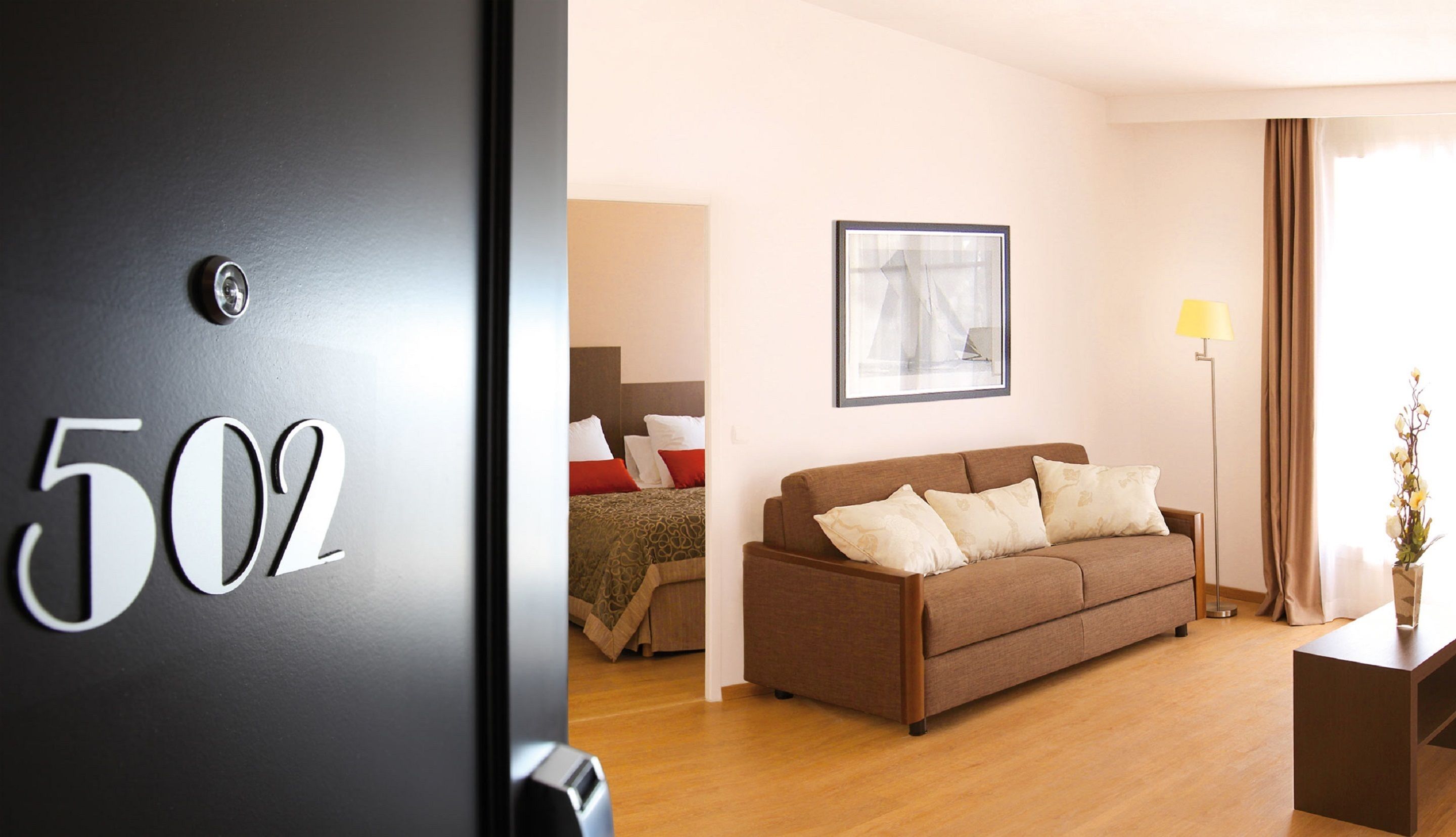A hotel room with a bed, a brown sofa, a framed picture on the wall, a floor lamp, and a door with the number 502.