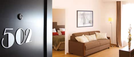 A hotel room with a bed, a brown sofa, a framed picture on the wall, a floor lamp, and a door with the number 502.