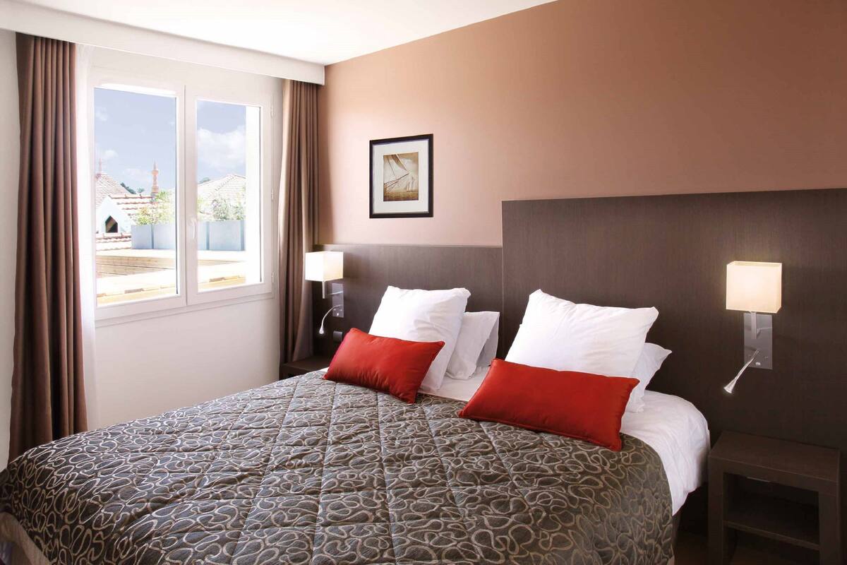 Una habitación de hotel con una cama grande, dos lámparas de noche, un cuadro en la pared y una ventana con cortinas.