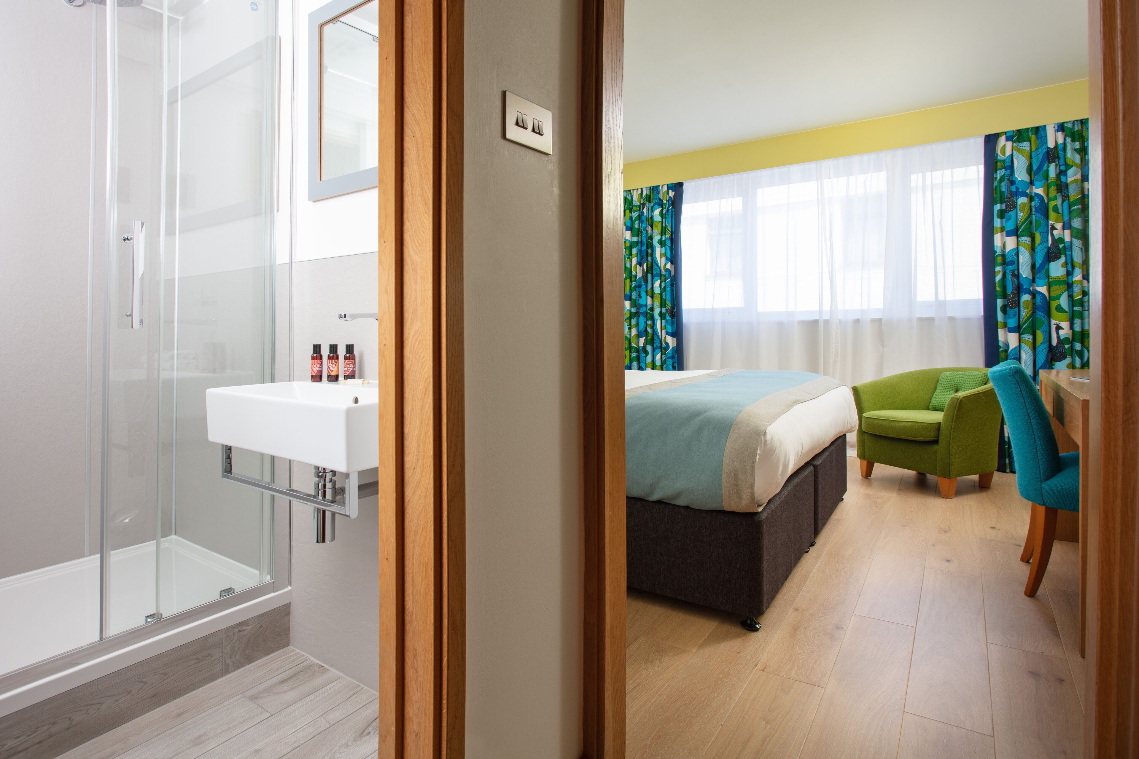 Une chambre d’hôtel moderne dotée d’une douche à parois de verre, d’un lavabo blanc, d’un lit avec un fauteuil vert et d’une fenêtre avec des rideaux.