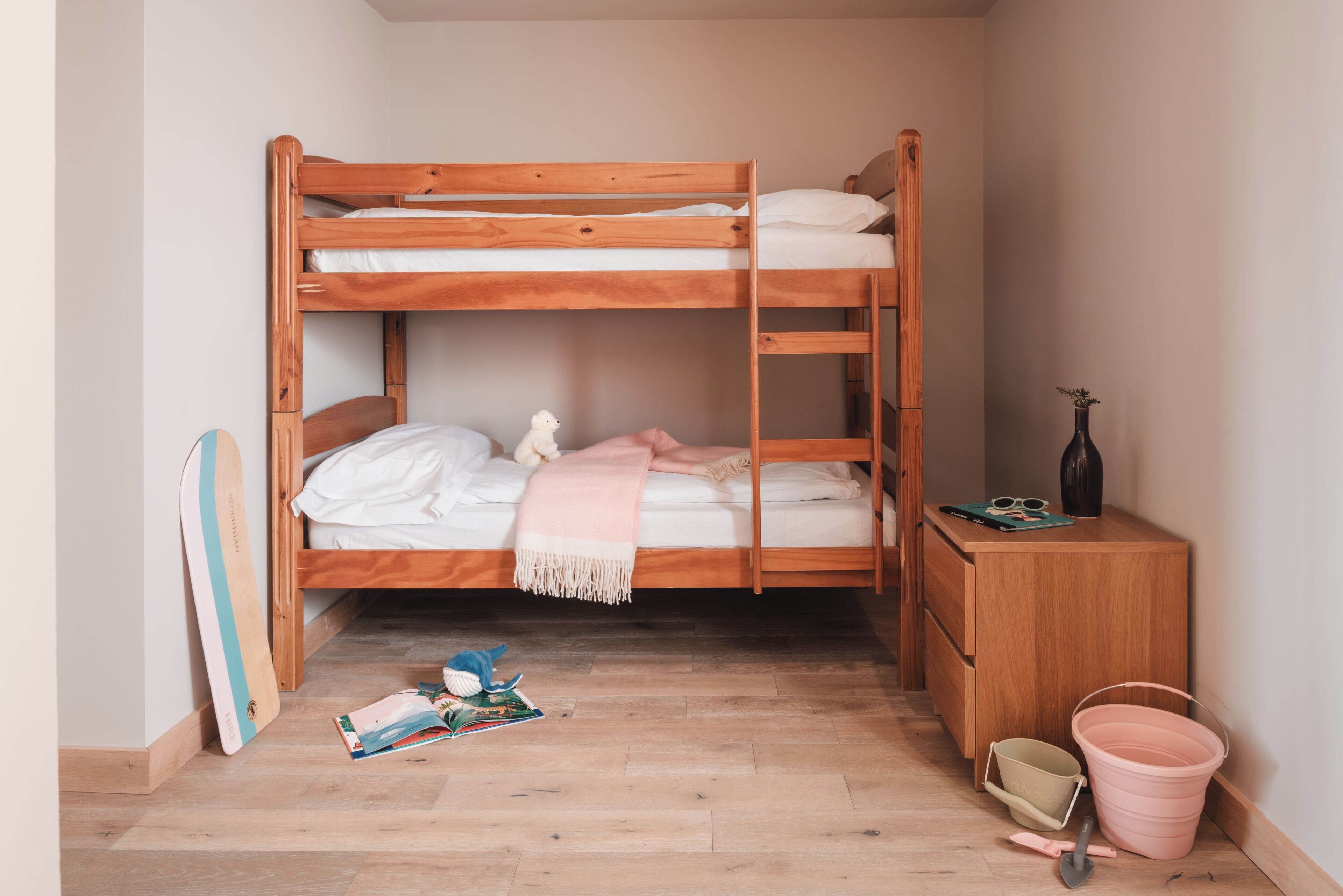 A bunk bed with a wooden frame, a small bedside table with a vase, and a pink bucket on the floor.