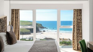 A bedroom with a large bed, a nightstand, a lamp, and a view of the beach and ocean through floor-to-ceiling windows. - Bedruthan Hotel and Spa (Newquay)