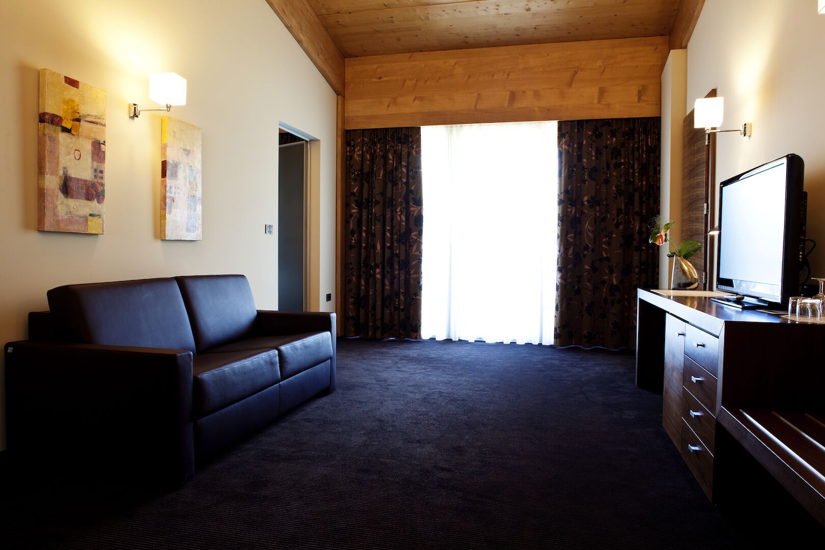 Une chambre d’hôtel avec une moquette sombre, un canapé en cuir, un bureau avec un téléviseur à écran plat et deux lampes fixées au mur.