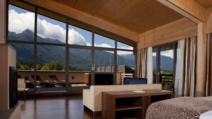 Habitación de hotel con un ventanal que ofrece vistas a la montaña, techo de madera, cama, escritorio y zona de estar con sillas.