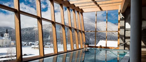 A glass-enclosed swimming pool with wooden beams and a view of a snowy village and mountains.