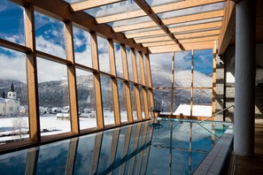 Une piscine intérieure, entourée de parois de verre, avec des poutres en bois et une vue sur un village enneigé et des montagnes.