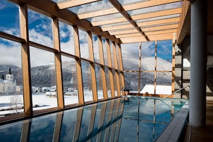 Una piscina rodeada de cristal con vigas de madera y vistas a un pueblo nevado y montañas.