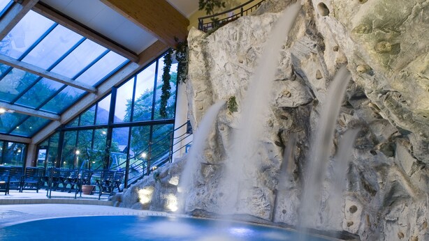 Una piscina interior con cascada, rodeada de paredes de piedra y vigas de madera.