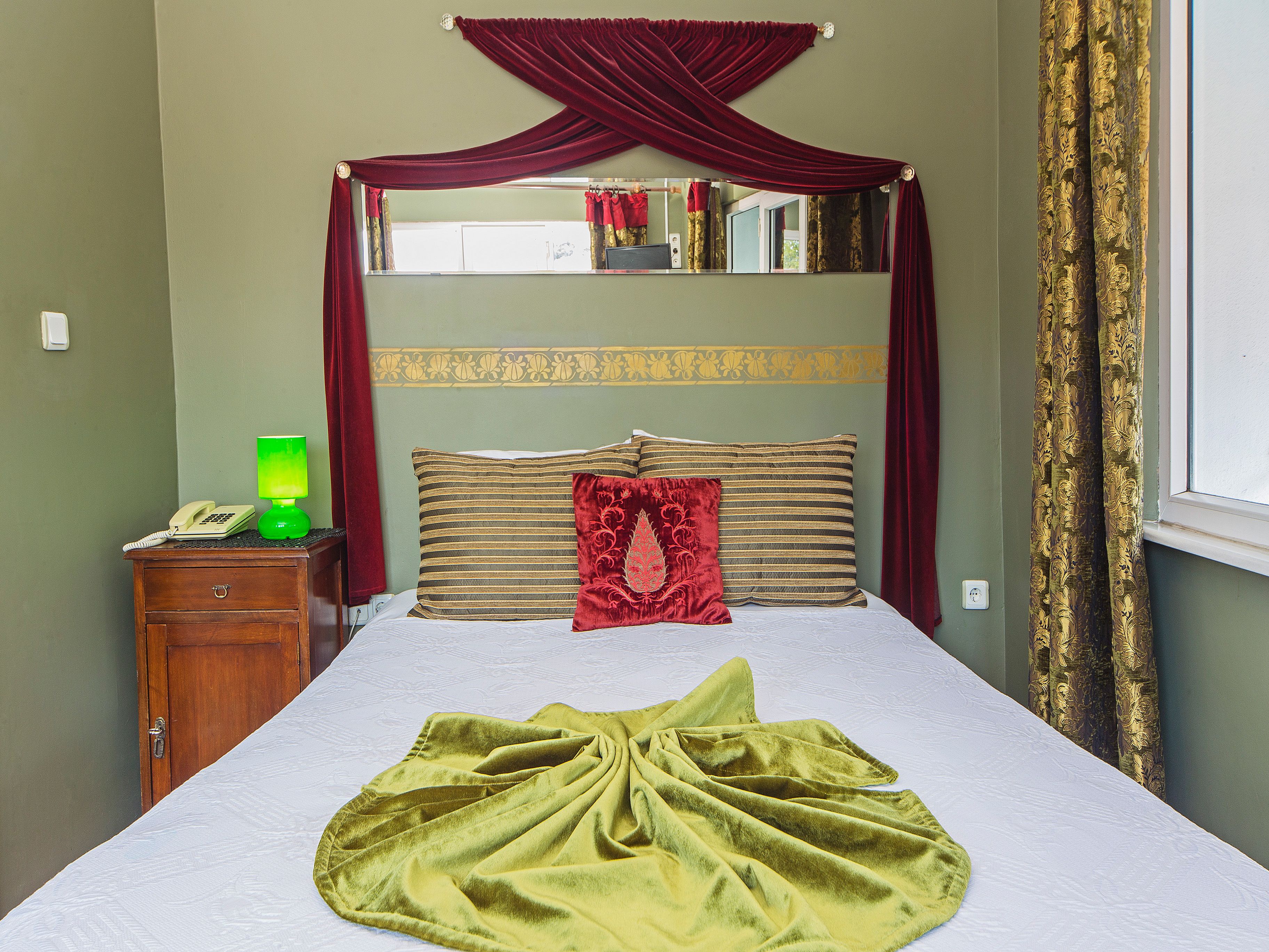 A neatly made bed with a green blanket, a wooden nightstand with a green lamp and a white telephone, and a mirror above the bed with a red curtain.
