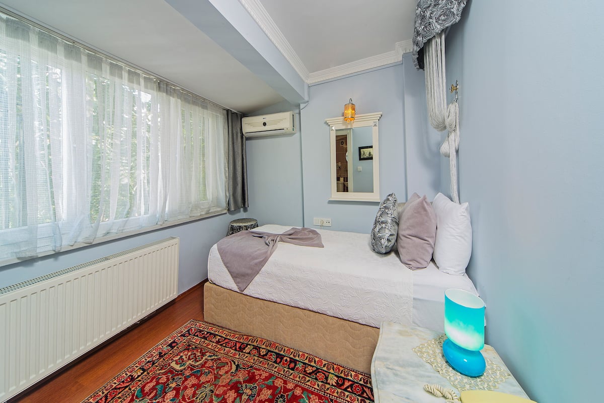 Une chambre à coucher avec un lit, une table de chevet avec une lampe, une fenêtre avec des rideaux légers, un radiateur et un tapis à motifs.