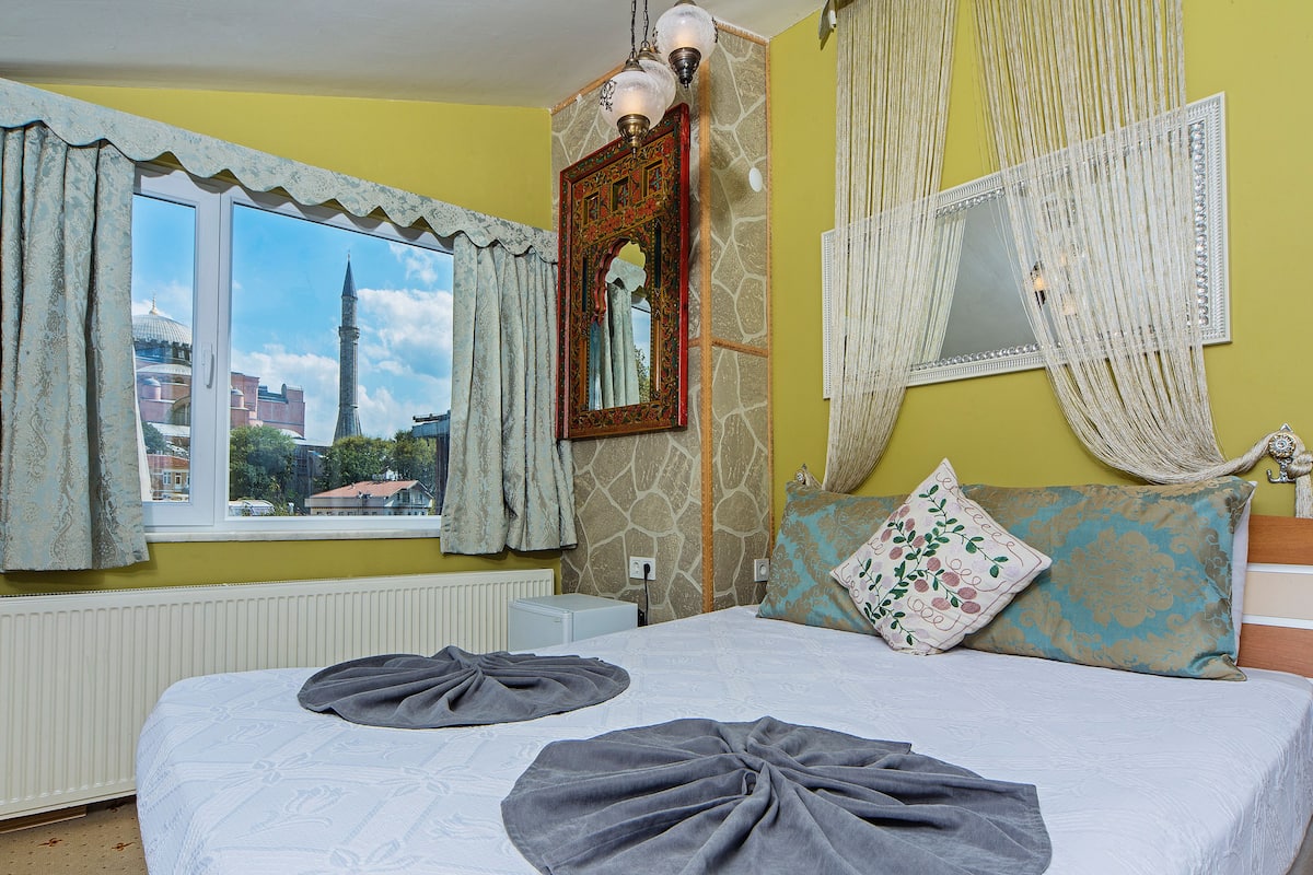 Une chambre à coucher avec un grand lit, une fenêtre avec des rideaux, un mur en pierre et une vue sur un bâtiment à l’extérieur.