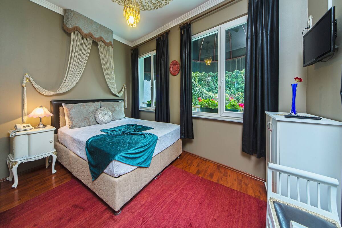 Une chambre à coucher avec un lit, une table de chevet, une lampe, une fenêtre avec des rideaux, un téléviseur et un vase avec une fleur.
