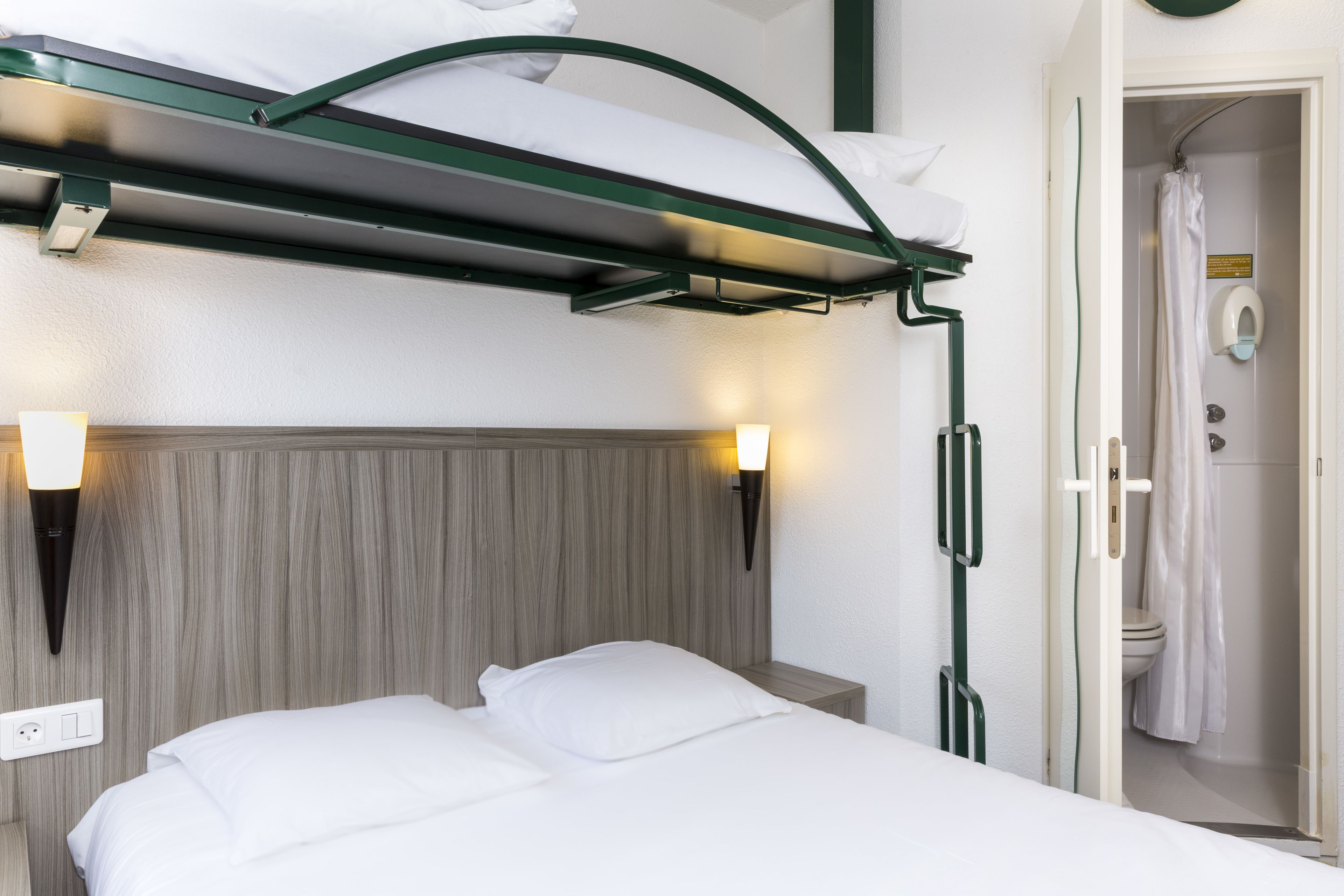 A hotel room with a bunk bed, bedside lamps, a wooden headboard, and a visible bathroom with a shower.