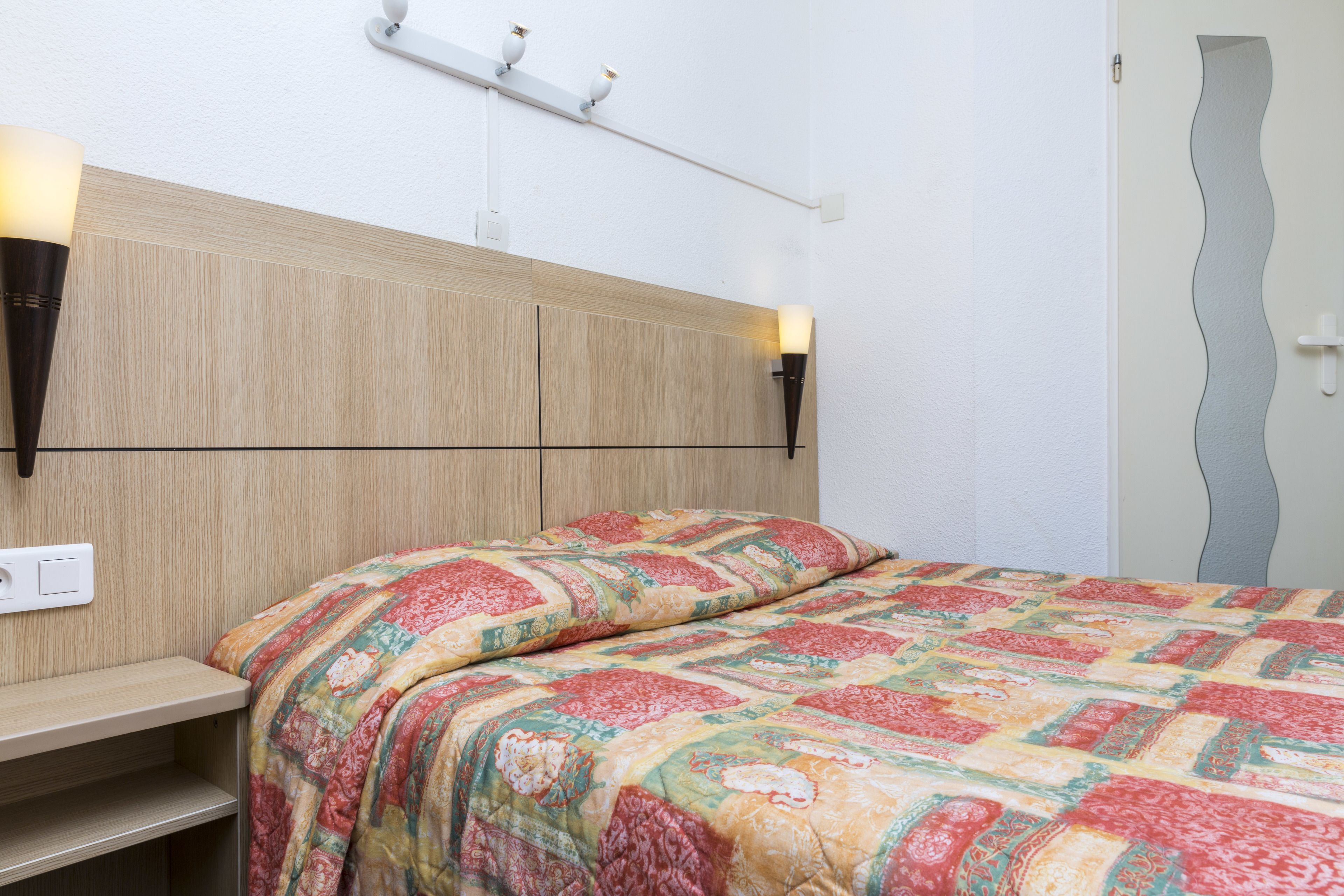 Un dormitorio con cabecera de madera, colcha acolchada, mesitas de noche e iluminación en la pared.