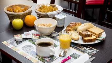 Una mesa de desayuno con un tazón de cereal, una naranja, una manzana, un croissant, tostadas, jugo, café y un periódico.