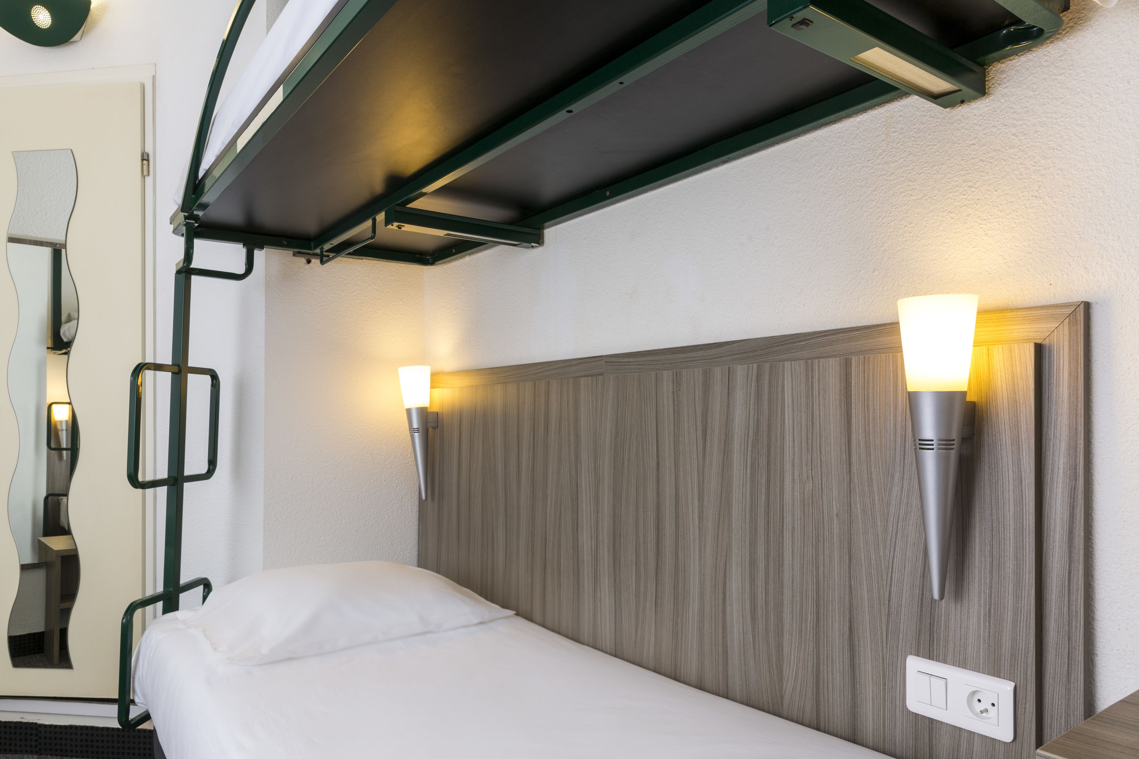 A hotel room with a bed, two wall-mounted lights, a power outlet, and a mirror with a unique design.