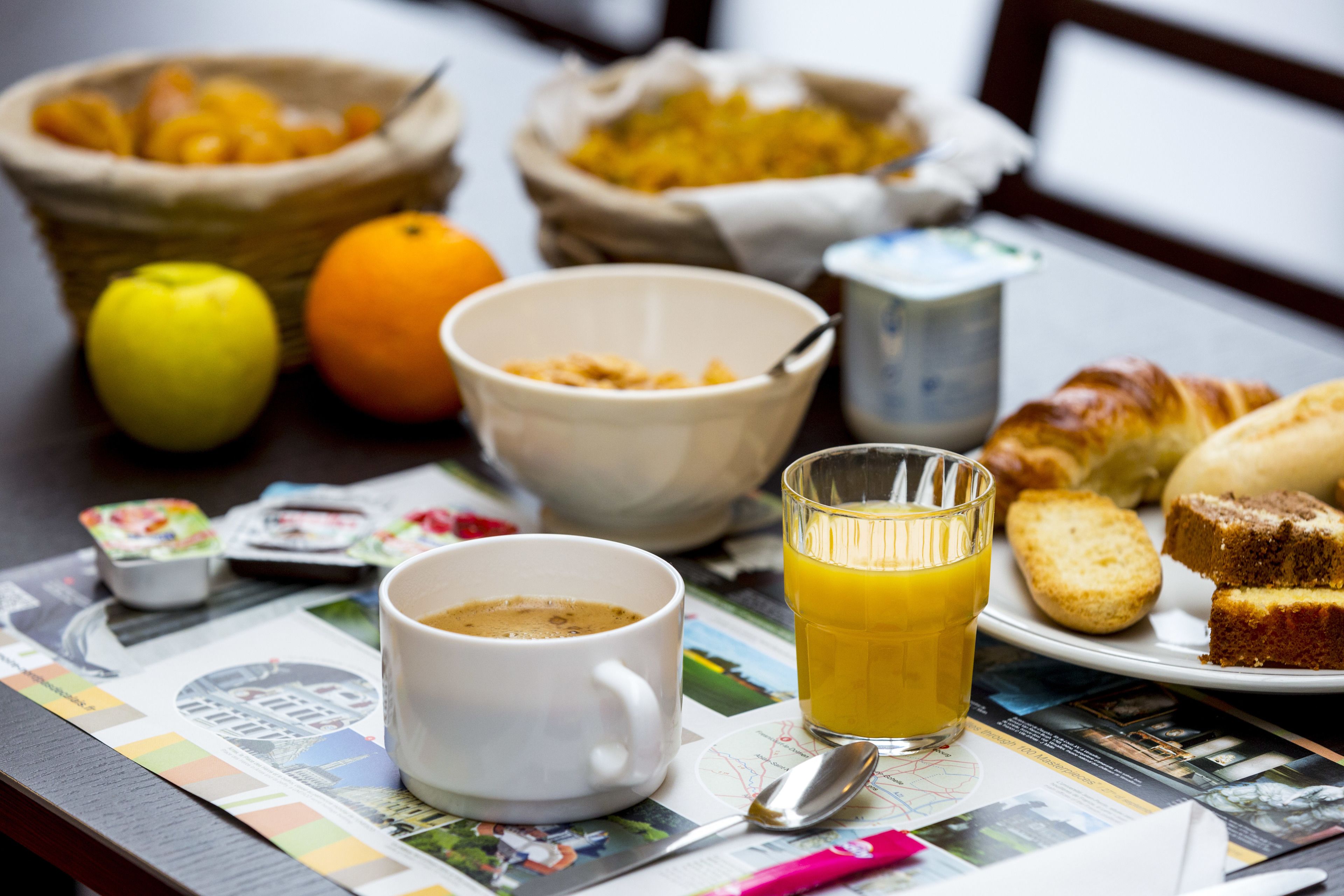 Una mesa de desayuno con un periódico, una taza de café, un vaso de jugo de naranja, un tazón de cereal, un croissant y una cesta de fruta.
