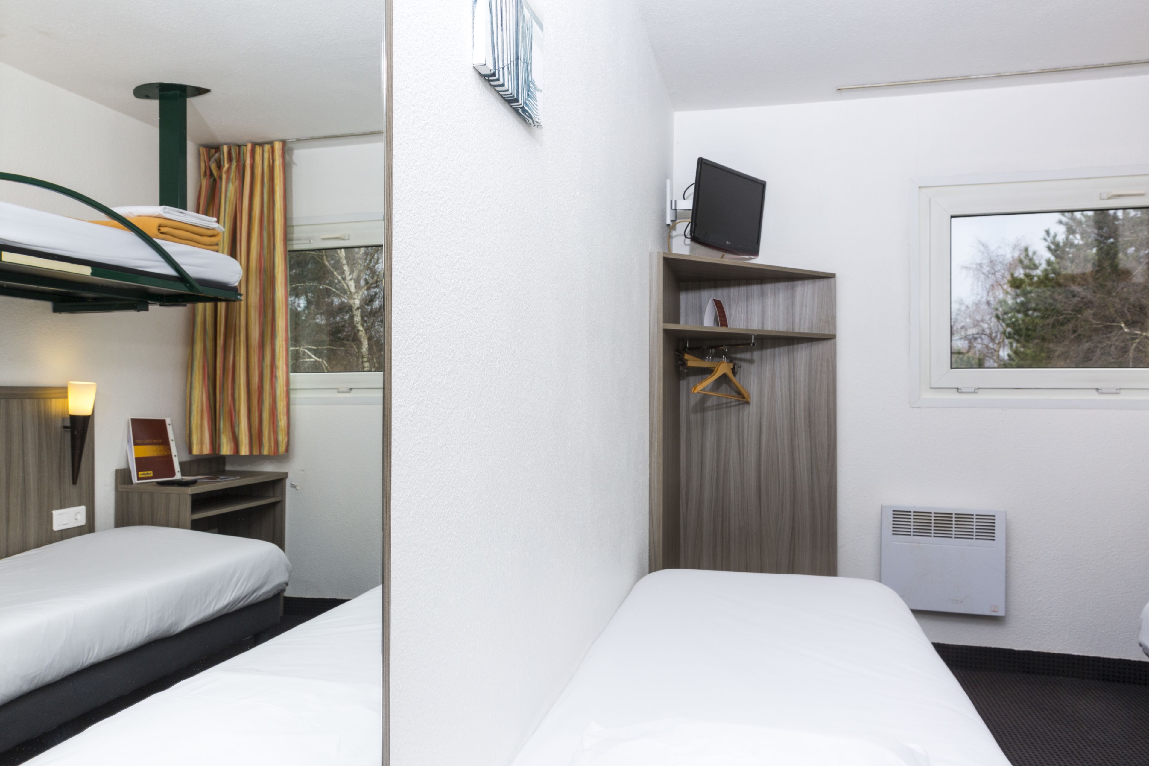 Ein Hotelzimmer mit zwei Betten, einem Fernseher, einer Heizung und einem Fenster mit Blick auf Bäume.