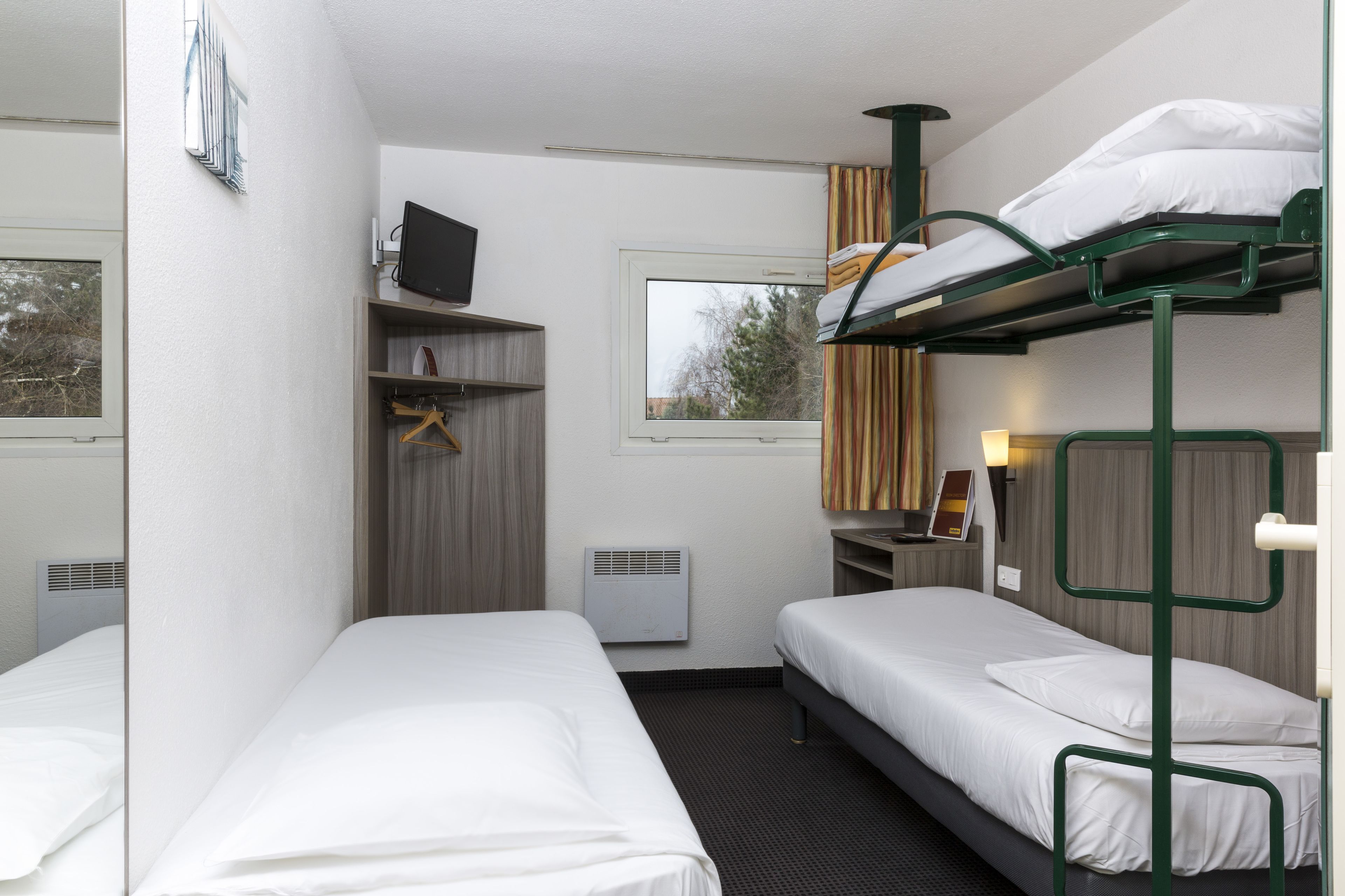 Habitación de hotel con dos camas individuales, una litera, un televisor, un calentador, un armario y una ventana con vistas.