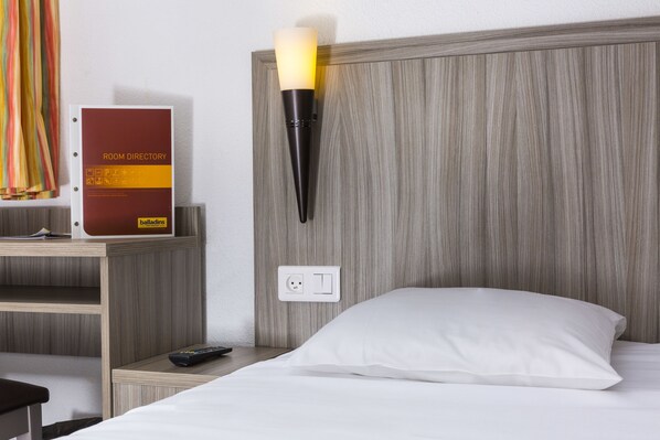 A hotel room with a wooden headboard, a bedside table with a remote control, a wall-mounted lamp, and a room directory sign.