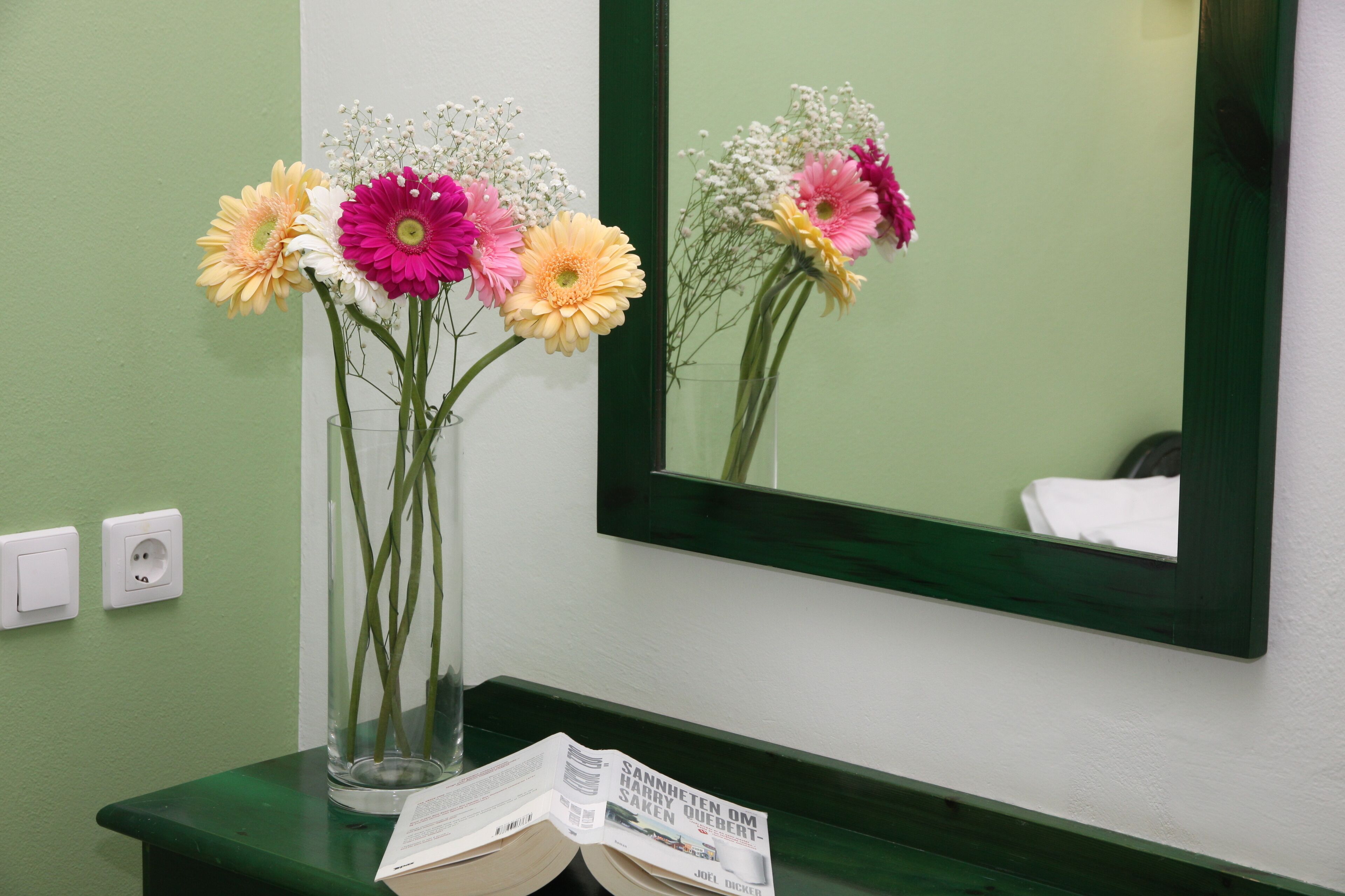 Um banheiro com bancada verde, espelho e um vaso com flores.