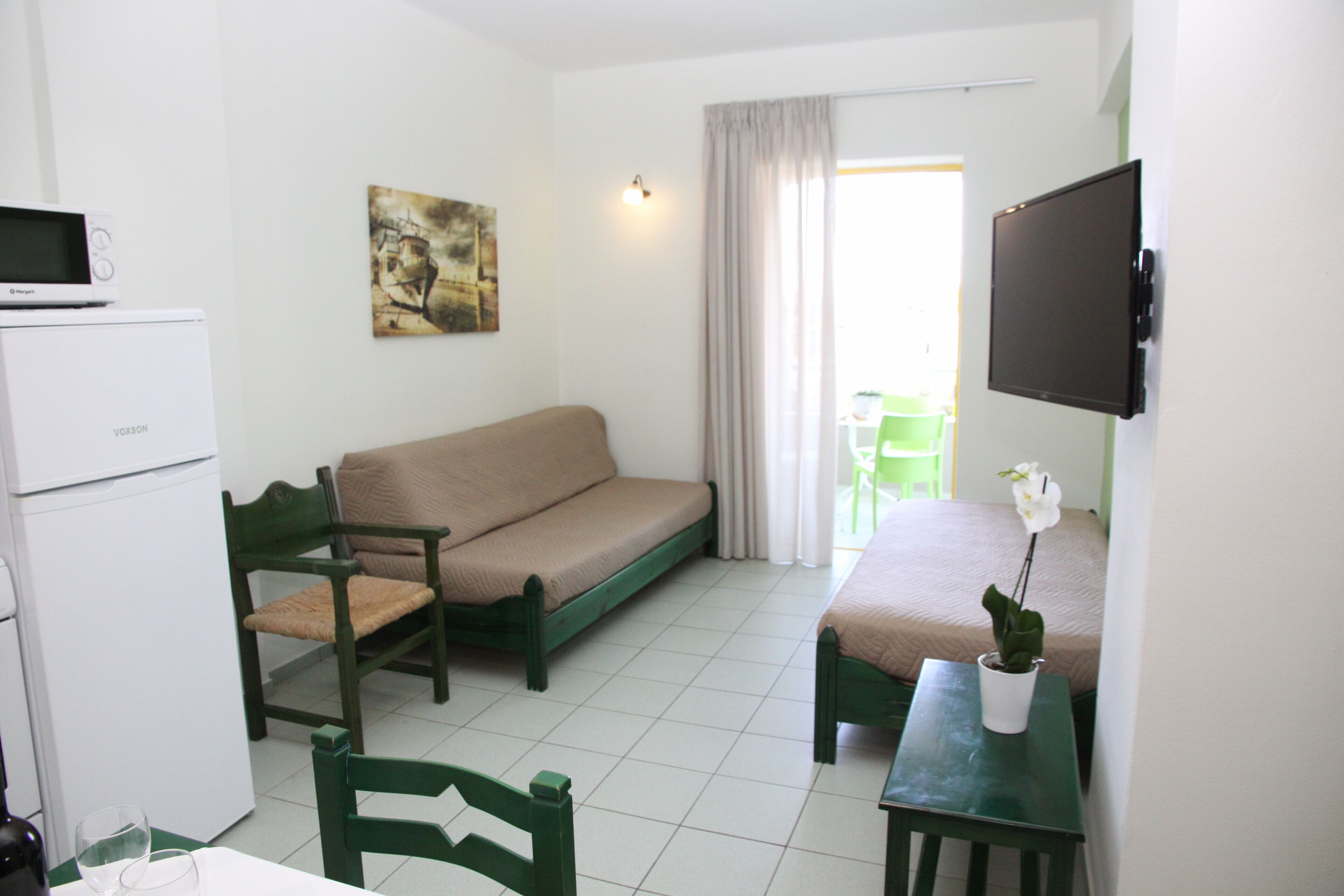 A small, clean room with a sofa, a chair, a small table with a potted plant, a flat-screen TV, and a refrigerator.
