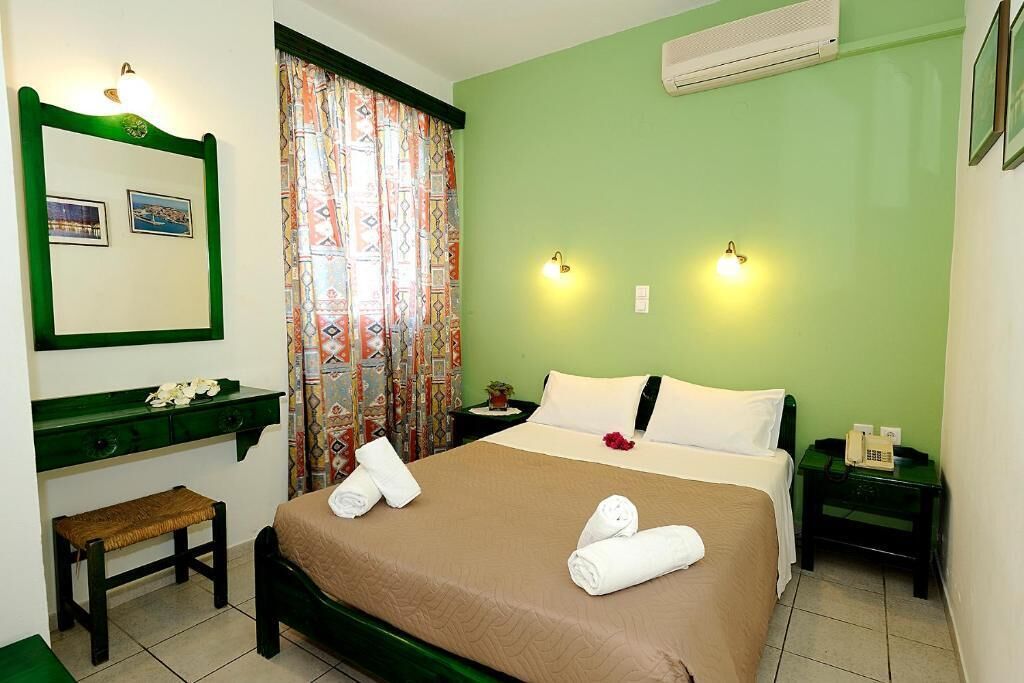 Habitación de hotel con cama, dos mesitas de noche, un banco, un espejo y una ventana con cortinas estampadas.