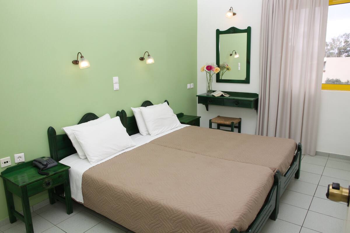 Una cama doble con cubrecama color beige, dos mesitas de noche con lámparas, una mesita con teléfono, un espejo sobre la cama y una ventana con cortinas.