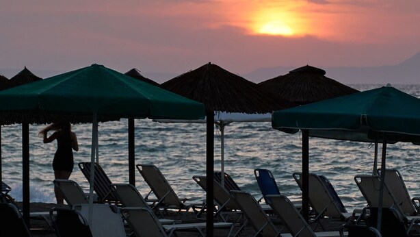A beach scene with lounge chairs and umbrellas, a person walking, and the ocean in the background. - Apollon Hotel Apartments (Rethymno)