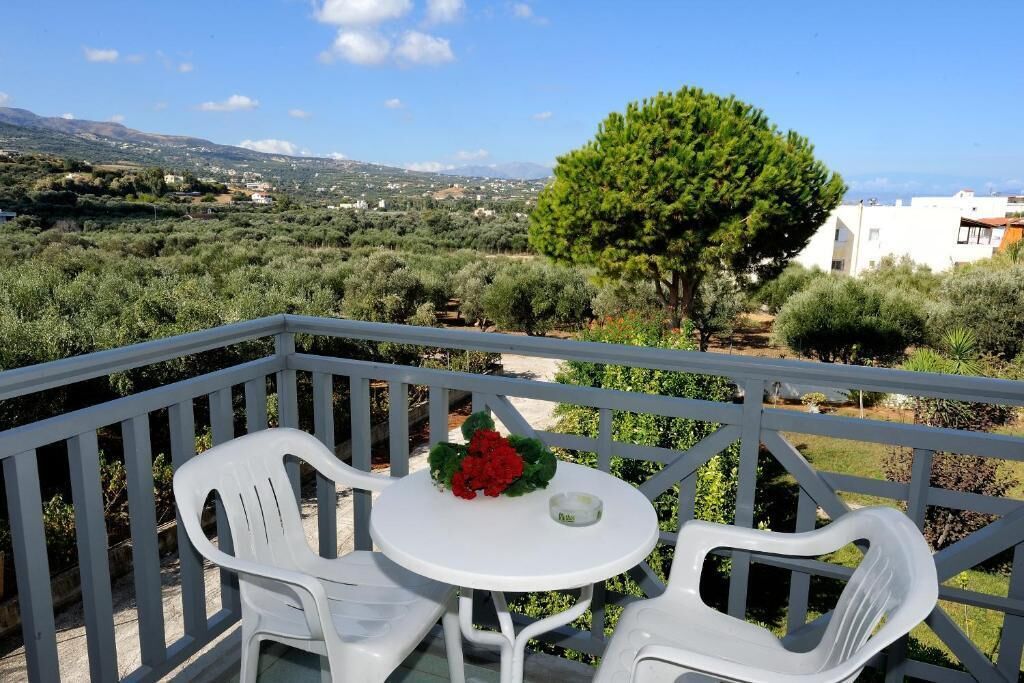 Un balcone con sedie di plastica bianca e un tavolino rotondo, che offre vista su uliveti ed edifici in lontananza.
