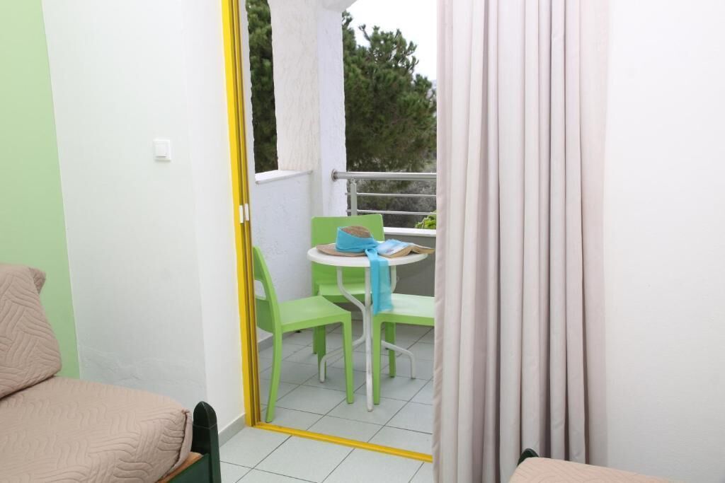 Ein Zimmer mit einem Bett, einem grünen Sessel, einem kleinen Tisch mit blauer Tischdecke und einem Balkon mit Blick auf Bäume.