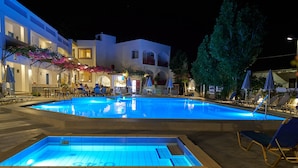 A swimming pool with lounge chairs and umbrellas, surrounded by buildings with outdoor seating and a night-time setting. - Apollon Hotel Apartments (Rethymno)