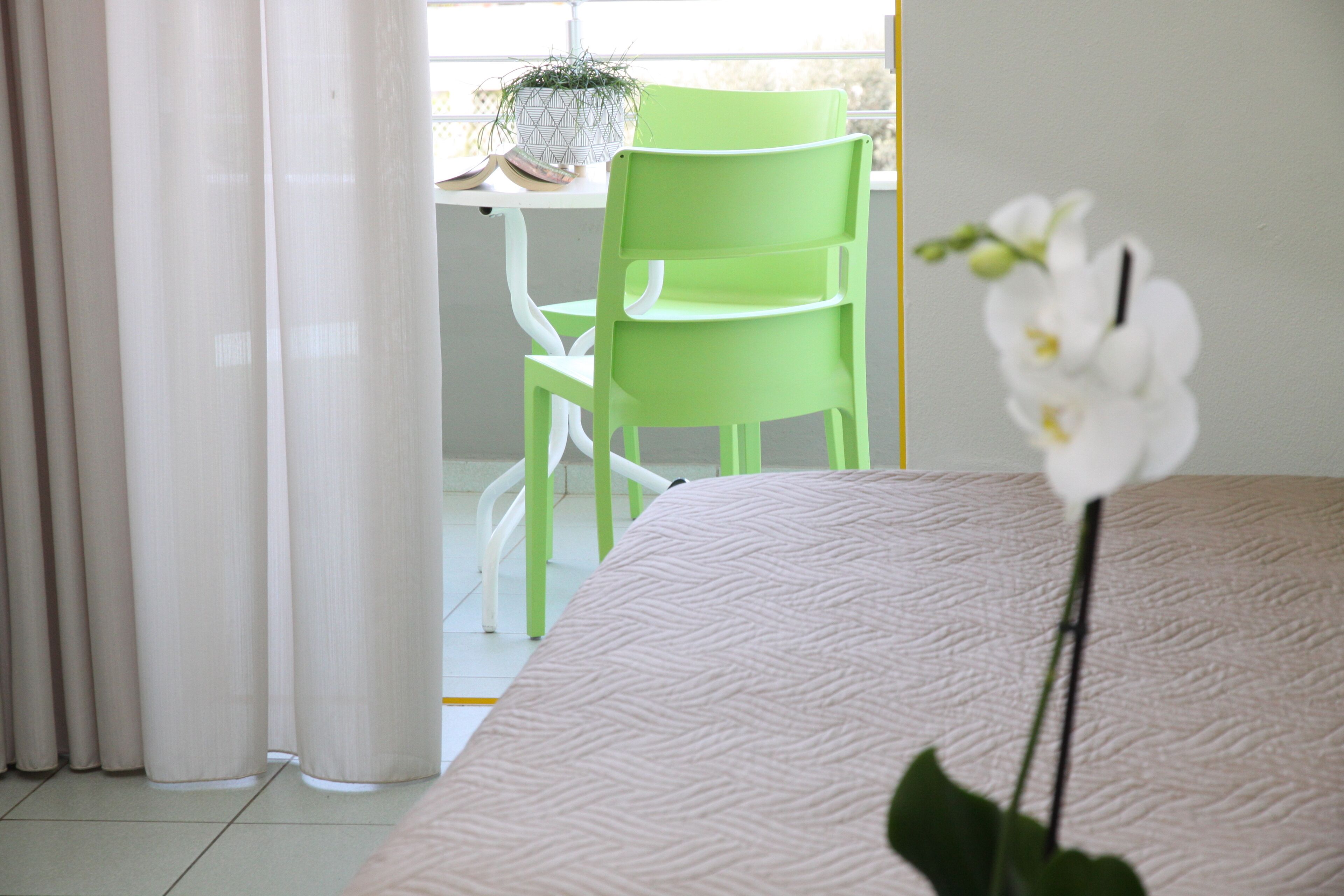 A room with a bed, a green chair, a table with a plant, and a window with curtains.